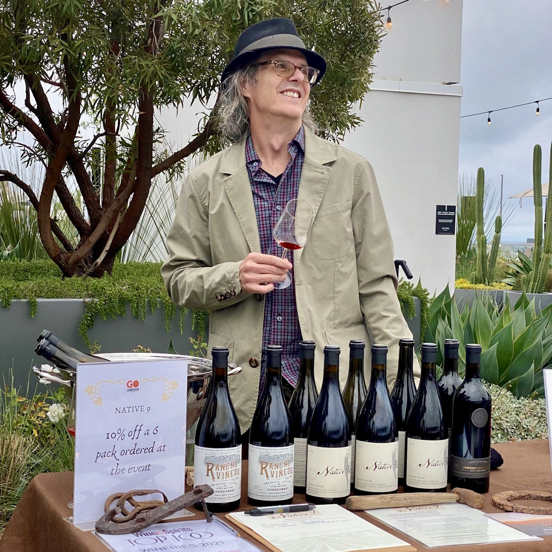 Wes smiles as he tastes wine at a table full of wine, wearing a fedora hat.