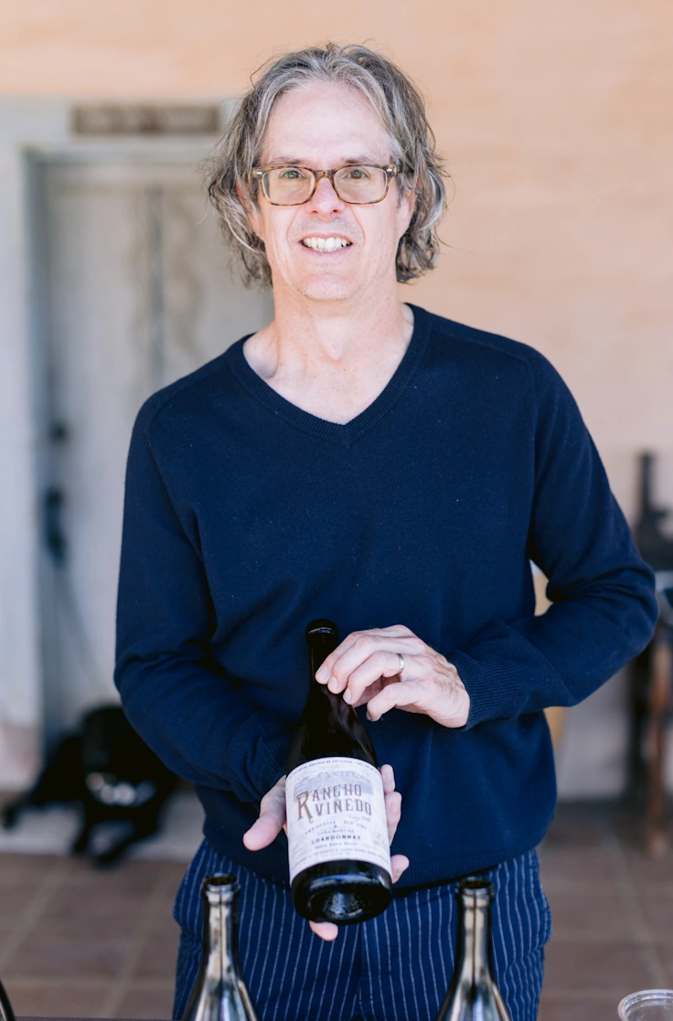 Wes Hagen, early 50's dude in a blue sweater and longish grey hair holds a bottle of wine.